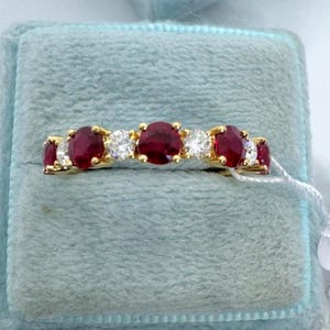 May include: A gold band ring featuring an alternating pattern of three round red gemstones and four round clear white gemstones set in a prong setting. The ring is displayed horizontally with a small white price tag attached to the side.