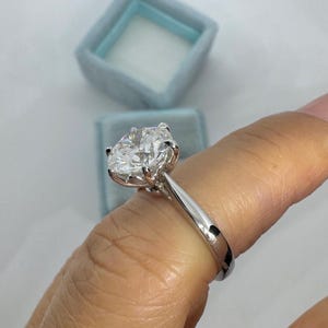 May include: A solitaire engagement ring with a large, clear, oval-cut diamond set in a silver band. The ring is displayed against a light blue ring box.