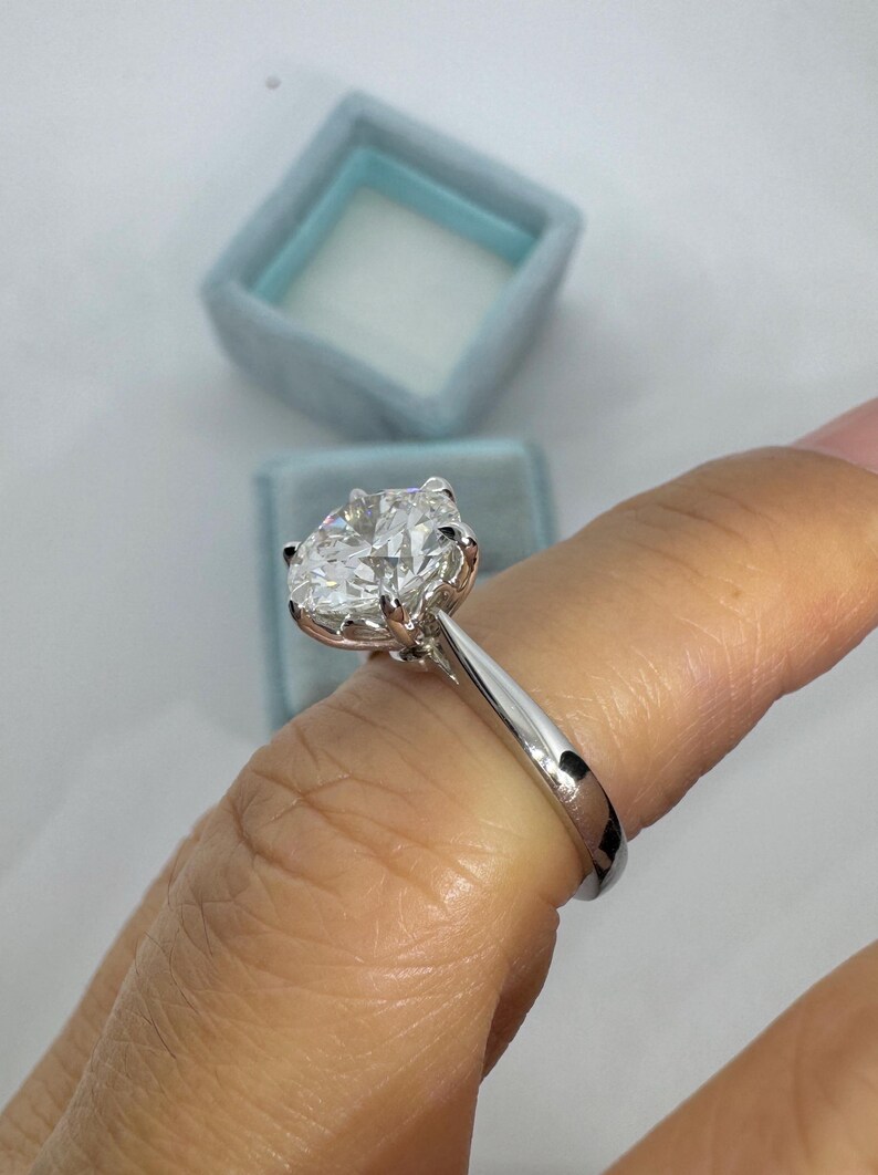 May include: A solitaire engagement ring with a large, clear, oval-cut diamond set in a silver band. The ring is displayed against a light blue ring box.