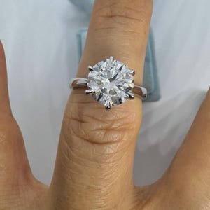 May include: A large, round-cut diamond ring with a six-prong setting. The ring is set on a silver band and is shown on a finger. The diamond sparkles brightly, reflecting light.