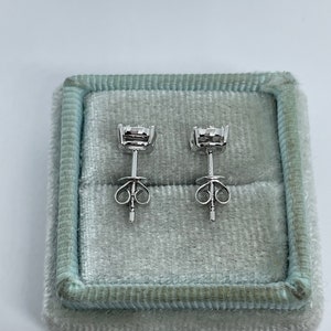 May include: A pair of silver stud earrings with small round clear stones set in a bezel setting. The earrings are in a light blue velvet box.