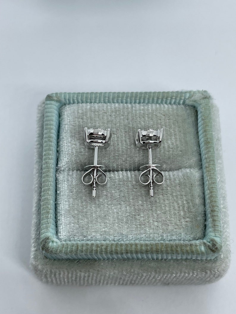 May include: A pair of silver stud earrings with small round clear stones set in a bezel setting. The earrings are in a light blue velvet box.