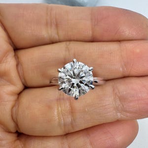 May include: A large, round-cut diamond ring with a six-prong setting. The diamond sparkles brilliantly, set on a simple, silver band. The ring is held in a person's hand, with a light blue jewelry box in the background.