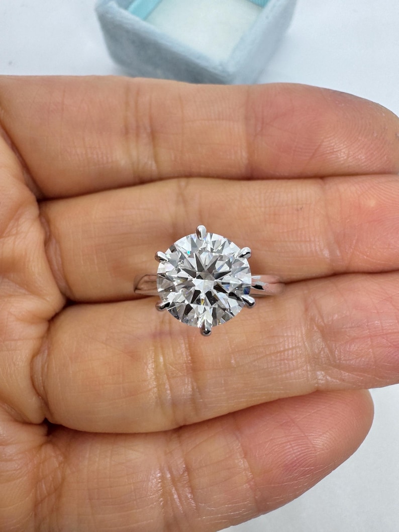 May include: A large, round-cut diamond ring with a six-prong setting. The diamond sparkles brilliantly, set on a simple, silver band. The ring is held in a person's hand, with a light blue jewelry box in the background.