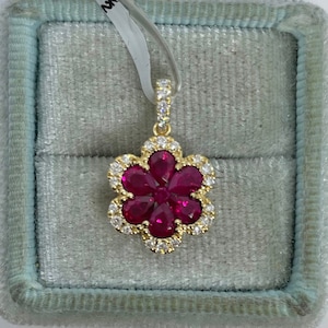 May include: A gold pendant in the shape of a flower, set with vibrant red gemstones at the center and surrounded by a halo of smaller, clear stones. The pendant is displayed in a light blue jewelry box.