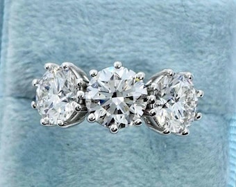 Könnte beinhalten: Ein Ring mit drei Steinen, mit einem großen, runden Diamanten im Brillantschliff in der Mitte, flankiert von zwei kleineren, oval geschliffenen Diamanten. Die Diamanten sind in einer Silber- oder Platinfassung gefasst, präsentiert in einer hellblauen Ringschatulle.