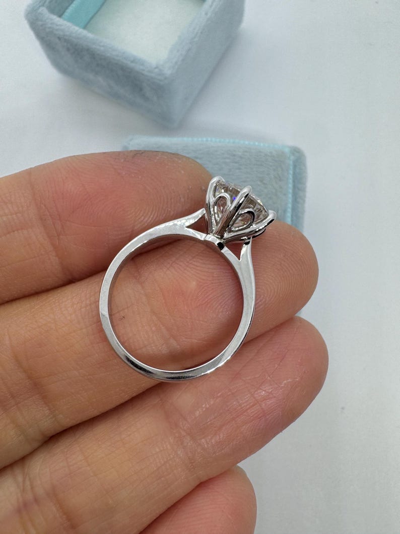 May include: A silver engagement ring with a large, clear gemstone. The ring has a classic solitaire design with a delicate band and a prong setting. A light blue ring box is in the background.