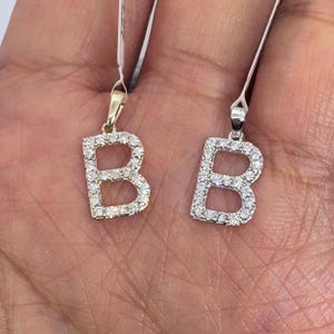 May include: Two letter "B" pendants, one in gold and the other in silver, each encrusted with small, sparkling gemstones. The pendants are suspended from thin chains, showcasing the intricate details of the jewelry.