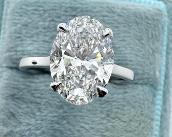 Könnte beinhalten: Ein Verlobungsring mit einem oval geschliffenen Diamanten. Der klare Diamant ist in einer silbernen Fassung mit vier Krappen gefasst. Der Ring liegt auf einem hellblauen Samthintergrund.