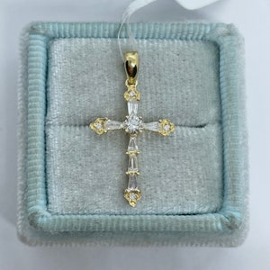May include: A gold pendant in the shape of a cross, set with clear gemstones. The cross is displayed inside a light blue velvet jewelry box. The pendant has a central round stone and baguette-cut stones.