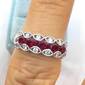 May include: A silver-toned ring featuring a central row of four round, deep red rubies. The rubies are flanked by two rows of small, clear, round-cut diamonds set within a decorative, milgrain-edged marquise-shaped metal framework.
