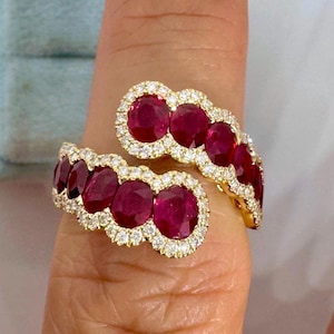May include: A bypass style ring crafted from yellow gold, featuring two rows of oval-cut deep red rubies. Each ruby is individually surrounded by a halo of small, round, brilliant-cut white diamonds, creating a continuous sparkling border.