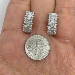 May include: A pair of silver hoop earrings with a row of baguette-shaped diamonds. The earrings are shown next to a US dime for size comparison.