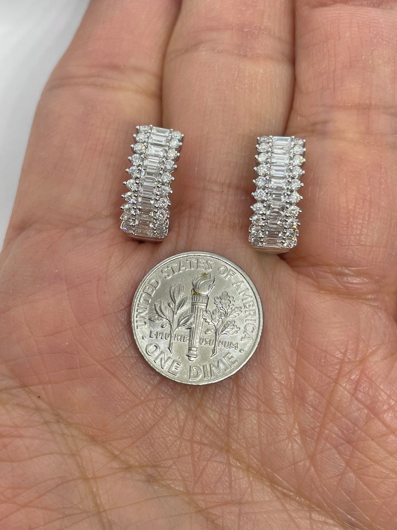 May include: A pair of silver hoop earrings with a row of baguette-shaped diamonds. The earrings are shown next to a US dime for size comparison.
