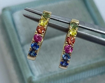 May include: A pair of gold hoop earrings, each featuring a row of colourful gemstones in a rainbow spectrum. The gemstones include blue, pink, orange, yellow, and green hues. The earrings are held by tweezers against a light blue background.