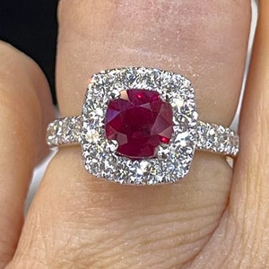May include: A ring with a large, red ruby gemstone surrounded by a halo of smaller diamonds. The ring is set in white gold and has a simple band.