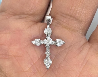 May include: A silver-colored pendant in the shape of a cross, adorned with numerous small, sparkling gemstones. The gemstones are clustered at the ends of the cross and along the vertical and horizontal beams. The pendant is held against a light background.