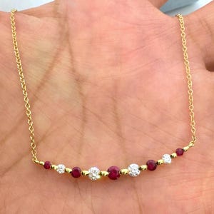 May include: A gold chain necklace featuring a curved bar design set with alternating round red rubies and clear white diamonds. The delicate metal chain connects to each end of the central gemstone arrangement, creating a balanced, horizontal jewelry piece.