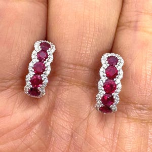 May include: A pair of hoop earrings featuring four round-cut deep red rubies set in a vertical row, bordered by a delicate frame of small, sparkling white diamonds set in polished silver-toned metal.