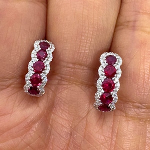 May include: A pair of hoop earrings featuring a row of four round, deep red rubies set in silver-toned metal, each surrounded by a halo of small, clear, sparkling white stones that create a scalloped edge along the length of the hoop design.
