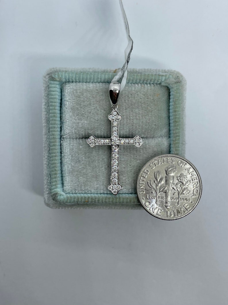 May include: A silver cross pendant with small clear stones set into the metal. The cross is in a blue velvet box next to a US dime for size comparison.