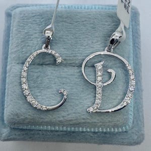 May include: Two silver pendants with diamond accents, one shaped like the letter C and the other shaped like the letter D, displayed on a light blue velvet background.