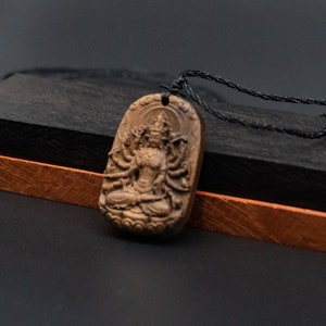 May include: A wooden pendant featuring a detailed carving of a multi-armed deity, with a black cord necklace. The pendant is resting on a black and brown wooden box.