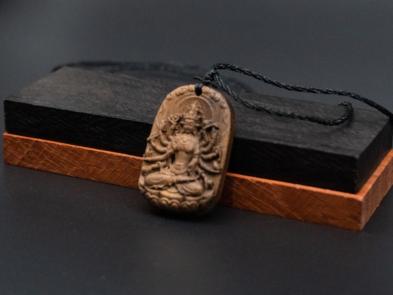 May include: A wooden pendant featuring a detailed carving of a multi-armed deity, with a black cord necklace. The pendant is resting on a black and brown wooden box.
