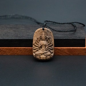 May include: A wooden pendant necklace featuring a detailed carving of a multi-armed deity. The pendant is brown and has a black cord.