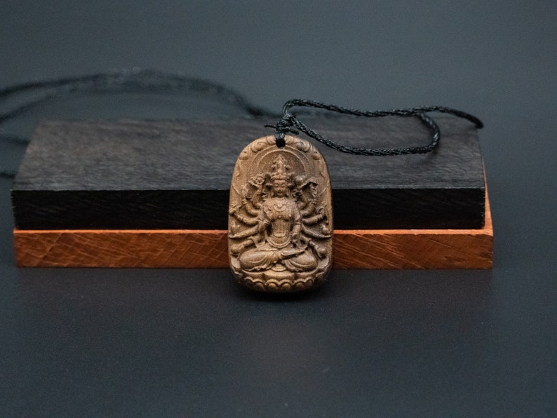 May include: A wooden pendant necklace featuring a detailed carving of a multi-armed deity. The pendant is brown and has a black cord.