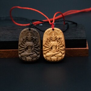 May include: Two wooden pendants with intricate carvings of a deity with multiple arms. The pendants are attached to a red and black cord.