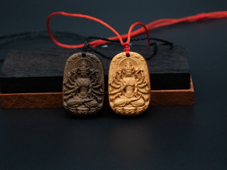 May include: Two wooden pendants with intricate carvings of a deity with multiple arms. The pendants are attached to a red and black cord.