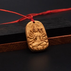 May include: A wooden pendant with a detailed carving of a deity with multiple arms. The pendant is attached to a red cord.