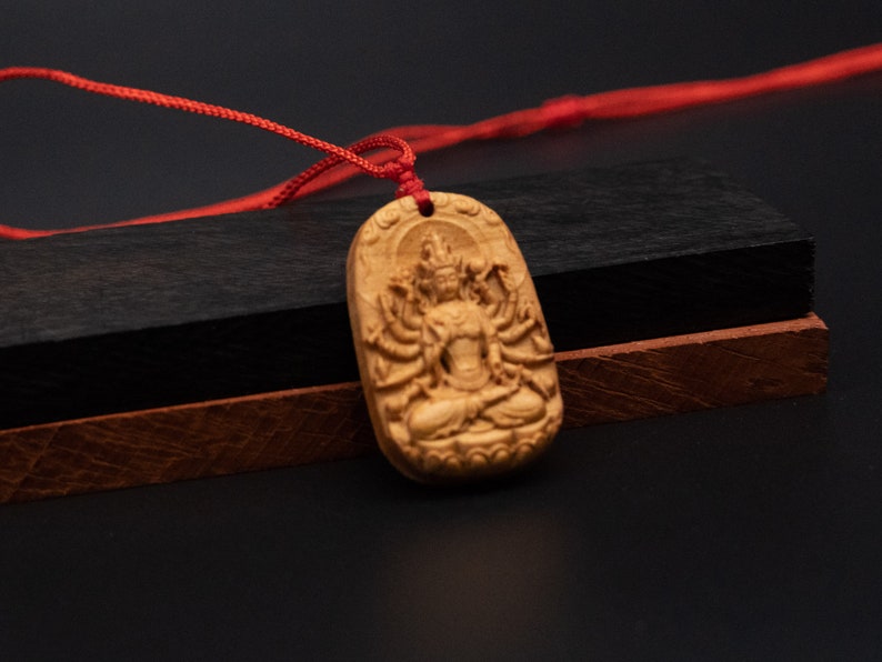 May include: A wooden pendant with a detailed carving of a deity with multiple arms. The pendant is attached to a red cord.