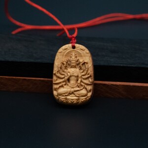 May include: A wooden pendant with a detailed carving of a deity with multiple arms, sitting in a meditative pose. The pendant is attached to a red cord.