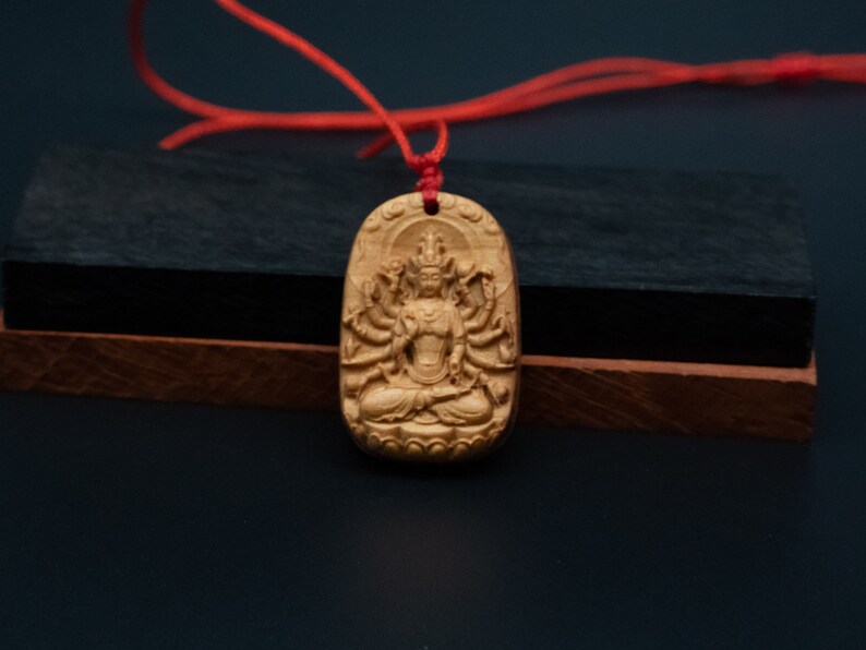 May include: A wooden pendant with a detailed carving of a deity with multiple arms, sitting in a meditative pose. The pendant is attached to a red cord.