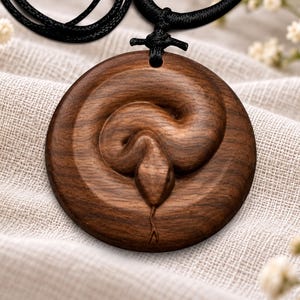 May include: A circular dark brown wooden pendant featuring a carved snake design in the centre. The snake is coiled in a spiral pattern with its head pointing downward and a visible forked tongue. The pendant hangs from a black cord necklace.