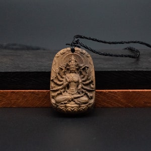 May include: A dark brown wooden pendant with a detailed carving of a deity with multiple arms. The pendant is hanging from a black cord.