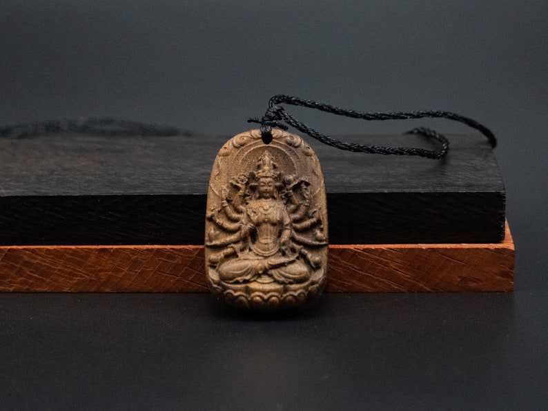 May include: A dark brown wooden pendant with a detailed carving of a deity with multiple arms. The pendant is hanging from a black cord.