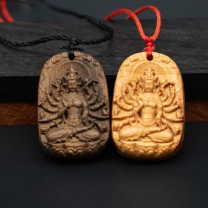 May include: Two wooden pendants with a carved image of a deity with multiple arms. The pendants are on black cords and are hanging against a dark background.