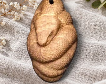 May include: An oval-shaped wooden pendant featuring a carved, coiled snake design with visible scale textures. The pendant is suspended from a black cord, showcasing natural light brown wood grain patterns throughout the surface.