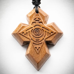 Può includere: Un ciondolo a forma di croce in legno marrone con un simbolo dell'occhio intagliato al centro. La croce ha una forma geometrica sfaccettata ed è appesa a un cordino nero. La venatura del legno è visibile sulla superficie del ciondolo, che presenta un design dettagliato e simmetrico con cerchi concentrici e motivi triangolari.
