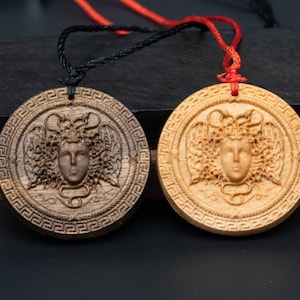 Könnte beinhalten: Zwei Holzketten mit einer geschnitzten Darstellung des Kopfes der Medusa. Eine Kette hat einen Anhänger aus dunklem Holz, die andere einen Anhänger aus hellem Holz.