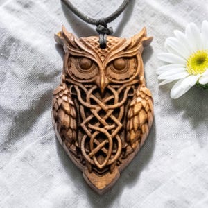 Puede incluir: Colgante de madera con forma de búho tallado con detalles intrincados, con un diseño de nudo celta. El colgante está suspendido de un cordón gris oscuro. Una margarita blanca con centro amarillo en el fondo. Mide unos 7 cm.