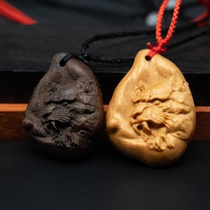Può includere: Due ciondoli in legno con volti di animali intagliati. Un ciondolo è marrone scuro e l'altro è marrone chiaro. Entrambi i ciondoli sono attaccati a cordini neri.