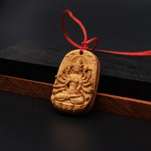 May include: A wooden pendant with a detailed carving of a deity with multiple arms, sitting in a meditative pose. The pendant is attached to a red cord.
