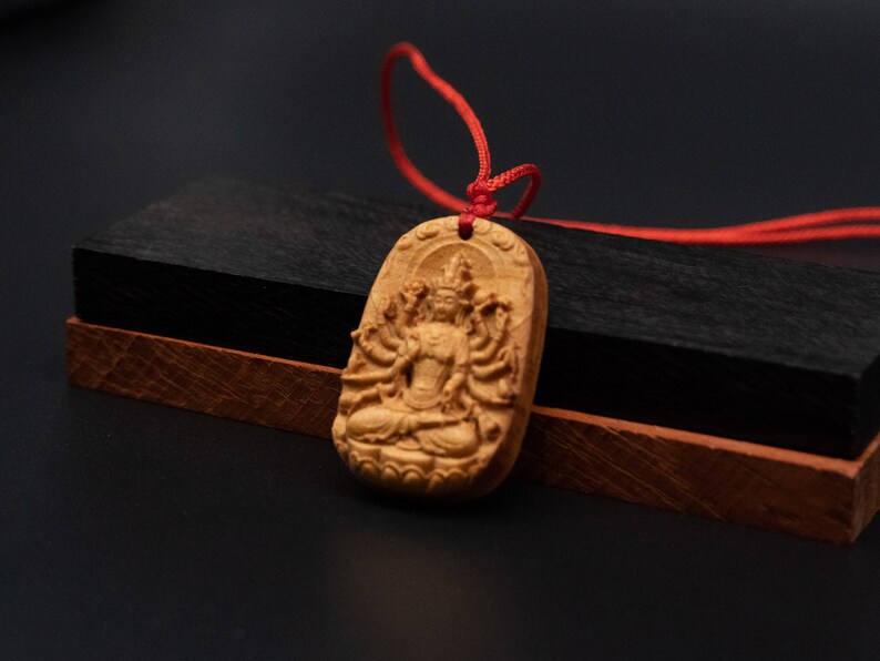 May include: A wooden pendant with a detailed carving of a deity with multiple arms, sitting in a meditative pose. The pendant is attached to a red cord.