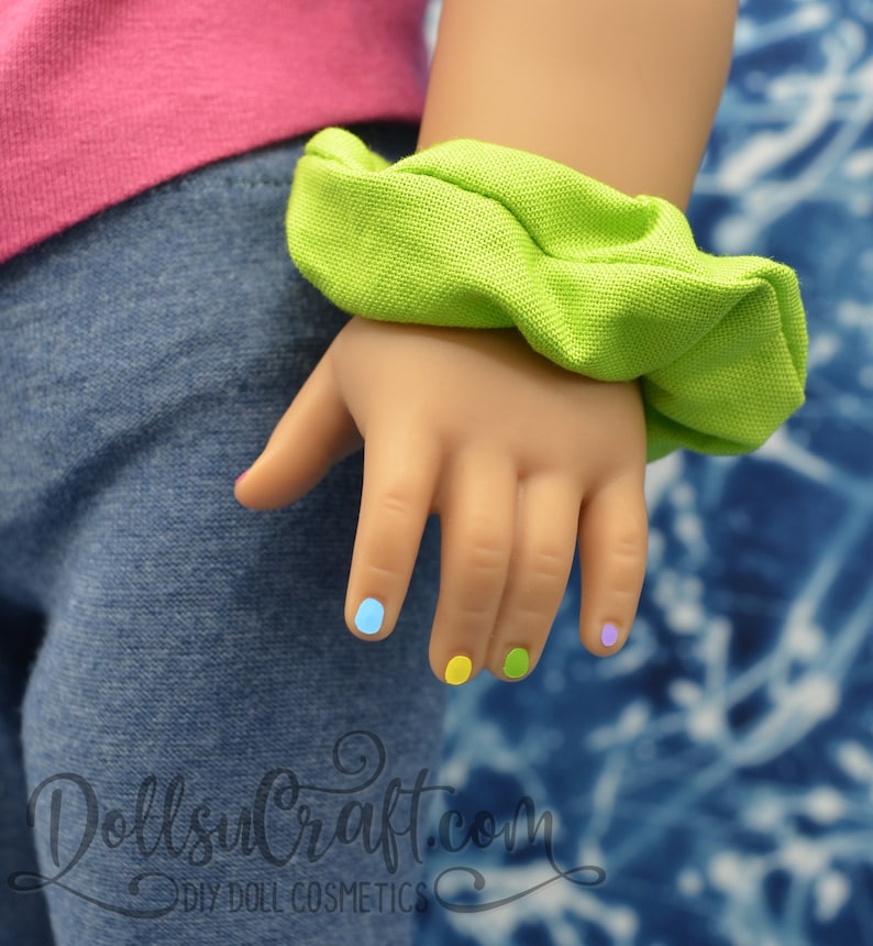 Puede incluir: Una mano de mu&ntilde;eca con esmalte de u&ntilde;as de colores. Las u&ntilde;as est&aacute;n pintadas en tonos de azul, amarillo y verde. La mano lleva una goma el&aacute;stica verde.