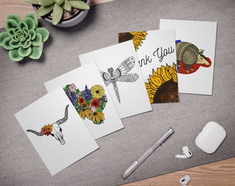 May include: Five blank greeting cards with different designs. The designs include a floral heart, a dragonfly, a sunflower, an armadillo, and a skull with flowers. The cards are on a gray surface with a pen, two clips, and two earbuds.