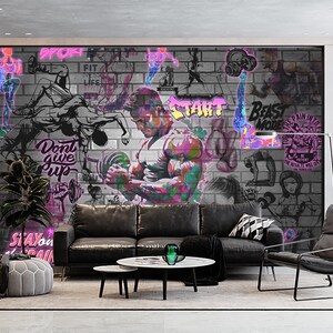 K&ouml;nnte beinhalten: Eine graue Backsteinwand mit bunten Graffiti, die motivierende S&auml;tze wie "Gib nicht auf", "Beast Mode" und "Start" zeigen. Die Wand ist mit Bildern von Menschen beim Sport verziert, darunter ein Mann, der Gewichte hebt. Die Wand befindet sich in einem Wohnzimmer mit einem schwarzen Ledersofa, einem schwarzen Ledersessel und einem Couchtisch.